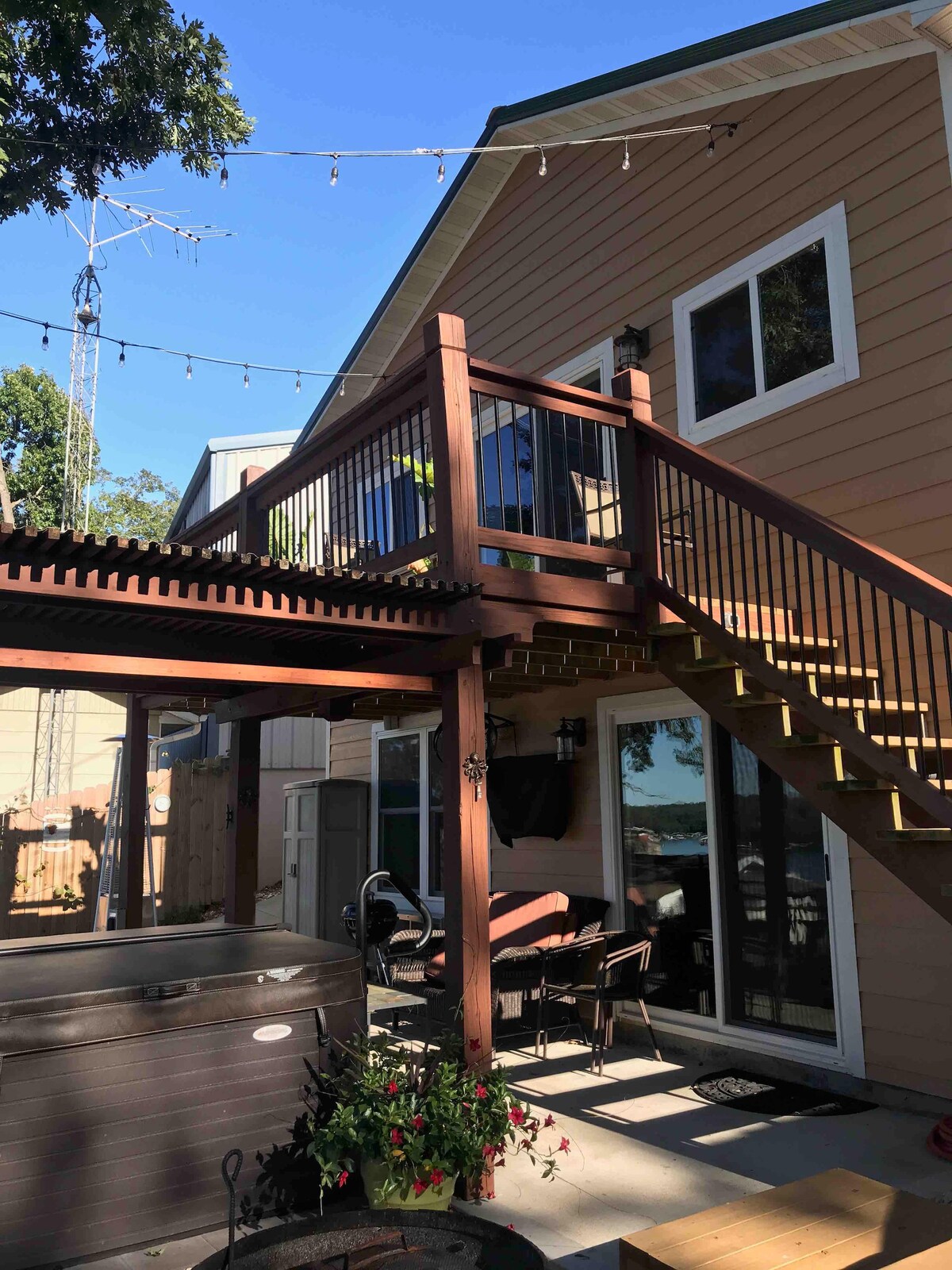 An outdoor space is shown featuring a wooden deck with stairs leading up to the guesthouse. A hot tub is visible on the lower level, alongside dining furniture. String lights are hung above the deck, creating an inviting atmosphere. The surrounding area includes greenery and soft natural lighting.