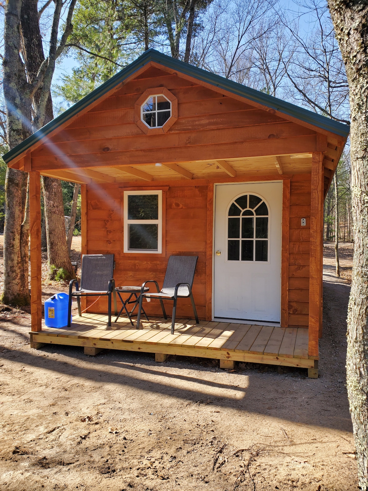 A rustic cabin exterior is shown, made of warm-toned wood. The front features a white door with a large window and a small circular window above it. Two chairs are placed on the wooden porch beside a blue container, set against a backdrop of trees.