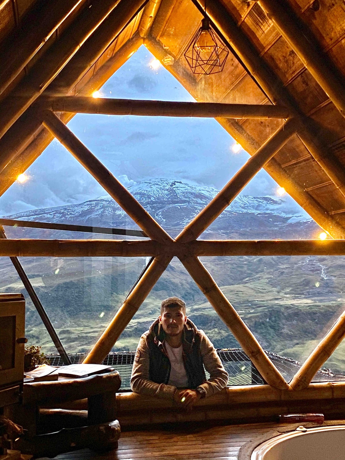 A striking view from the cabin features a large triangular window framing the Nevado del Ruiz mountain in the background. Soft lighting illuminates the wooden beams, creating a cozy ambiance. A seating area is positioned near the window, enhancing the picturesque scenery.