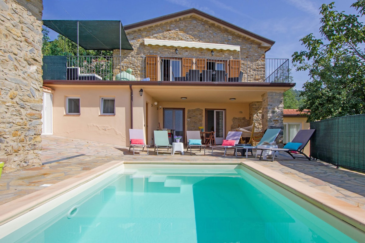 The exterior of the villa features a private pool and a spacious terrace lined with colorful loungers. The two-story stone structure is complemented by large balcony doors, providing access to the upper level. Lush greenery is visible in the background, enhancing the tranquil setting.