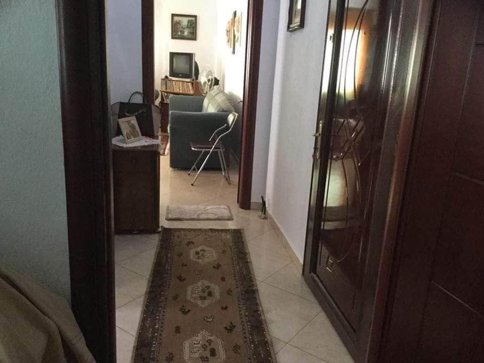 A hallway is visible, featuring a decorative runner carpet that extends towards a cozy sitting area. The space includes a small table with a framed picture and a chair. A closed door leads to the outside. Soft lighting adds to the inviting environment.