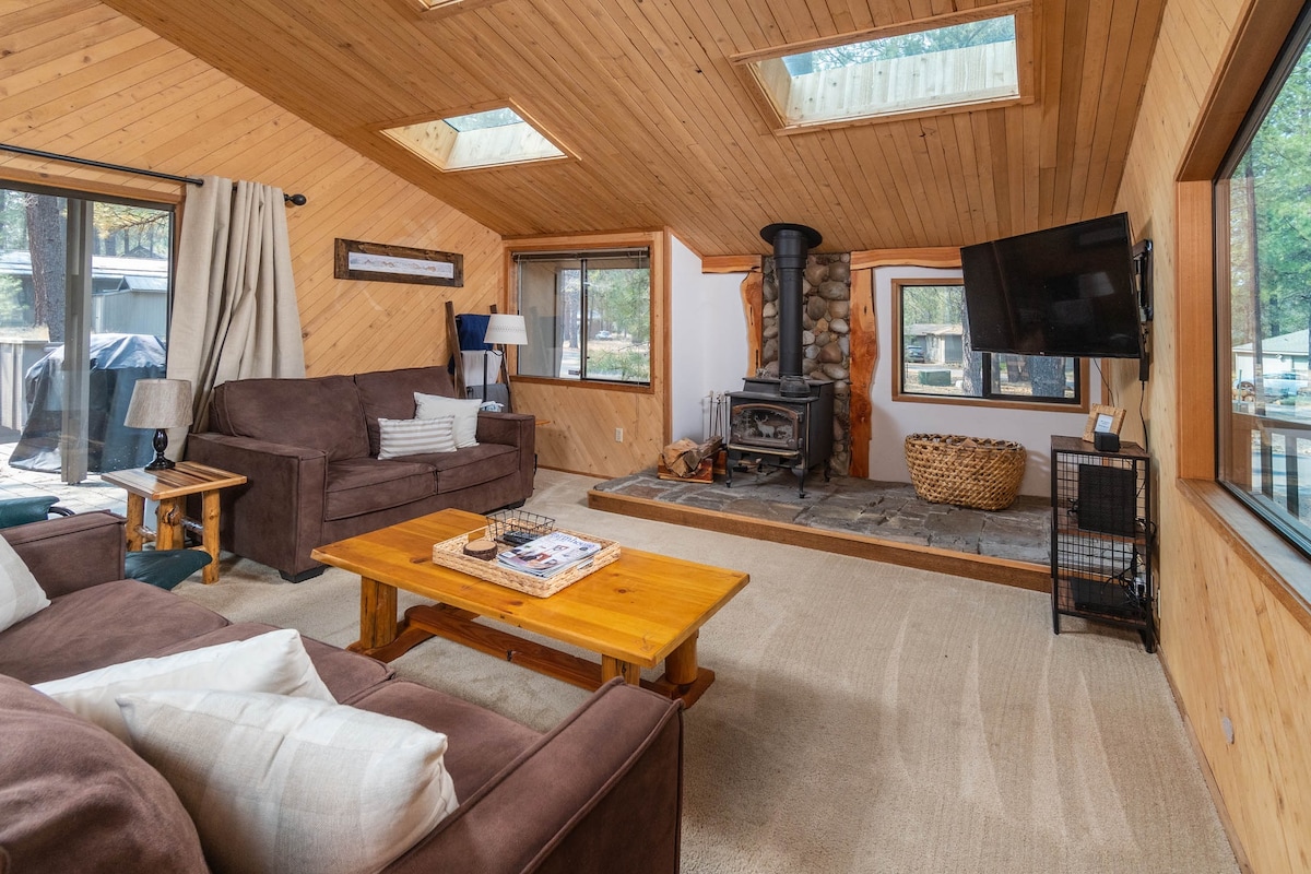 The cozy living area features a comfortable brown sofa and armchair arranged around a wooden coffee table. A stone fireplace is positioned centrally, with a large window allowing natural light to fill the space. Exposed wooden ceilings and walls create an inviting atmosphere.