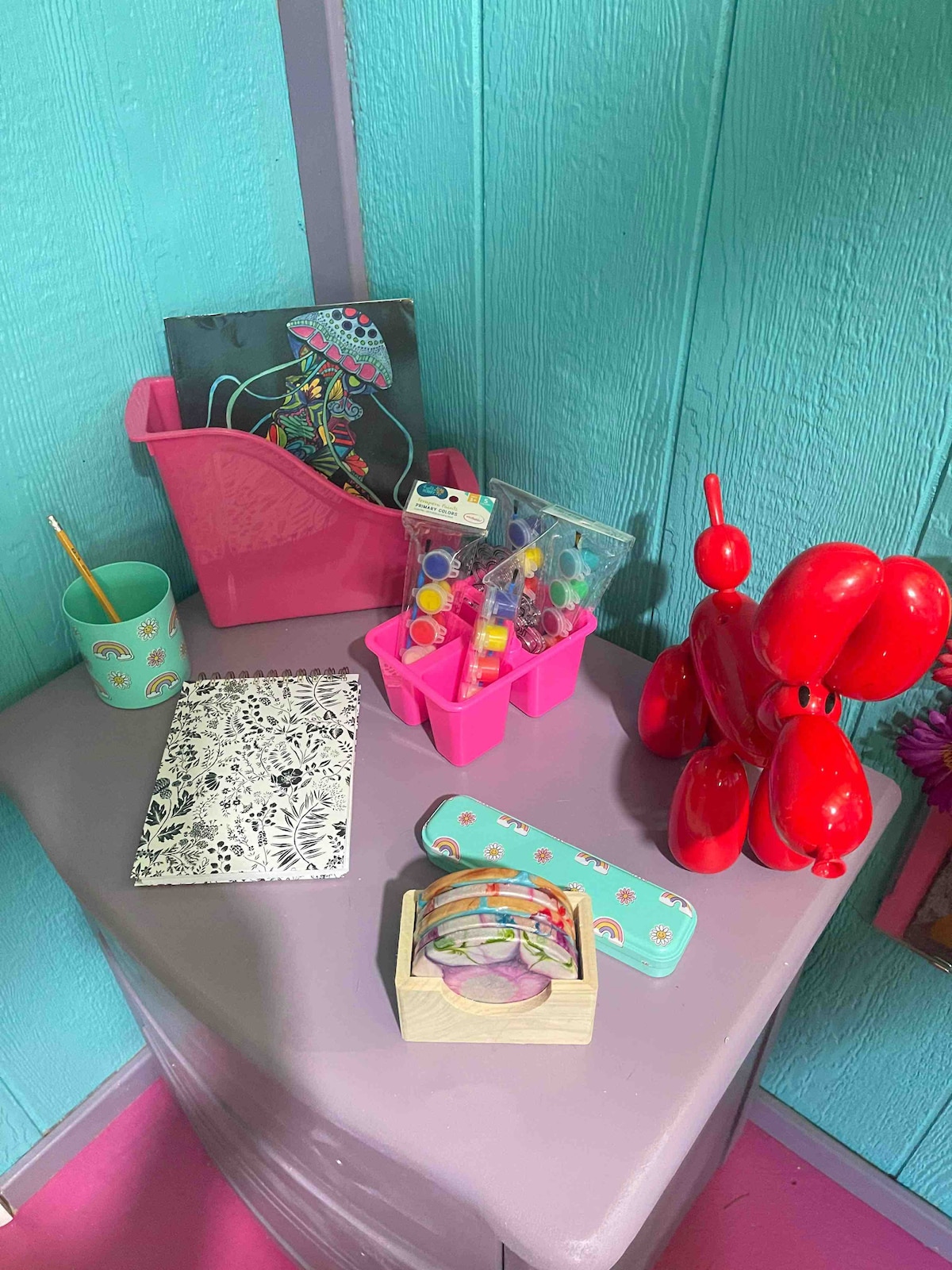 A colorful tabletop displays various playful items, including a pink storage caddy filled with craft supplies, a patterned notebook, and a whimsical balloon animal sculpture. Bright colors and a vibrant background enhance the creative atmosphere of the space.