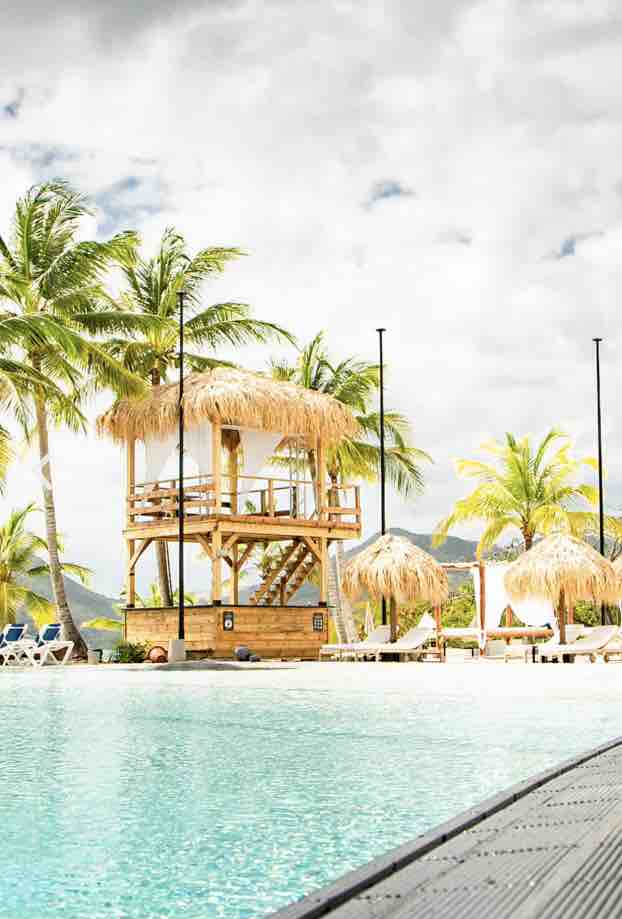 A wooden cabana with a thatched roof overlooks a large swimming pool surrounded by palm trees. Sun loungers with umbrellas are arranged nearby, creating a relaxing outdoor space under a partly cloudy sky.