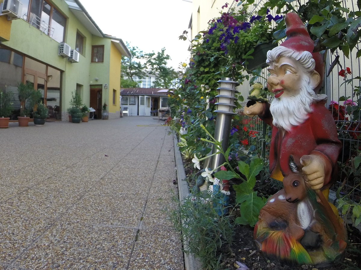 A decorative garden gnome is positioned amid vibrant flowers and greenery along a pebbled pathway. The surroundings feature a combination of potted plants and outdoor lighting, leading towards a spacious courtyard with adjoining buildings in the background.