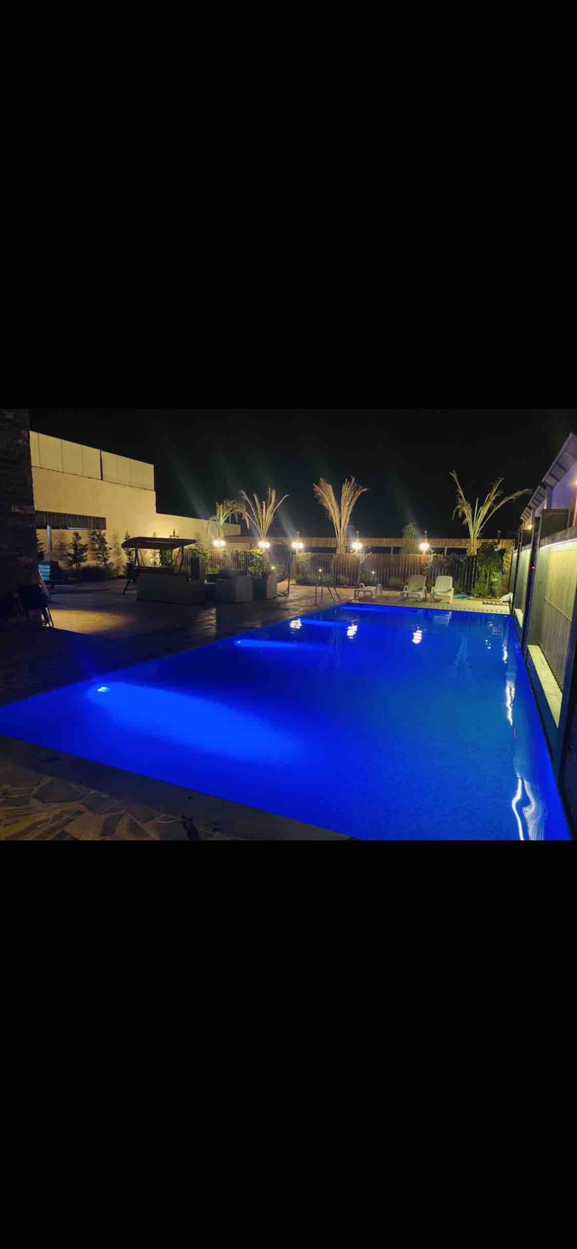 A large swimming pool is highlighted under the night sky, showcasing vibrant blue water illuminated by subtle lighting. Surrounding palm trees add greenery to the tranquil atmosphere, while lounge areas and landscaping can be seen in the background.