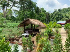 A traditional-style gazebo with a thatched roof is surrounded by a variety of potted plants. A circular table and chairs are visible inside the gazebo. The pathway leading to it is lined with greenery, while the backdrop features lush trees and hills.