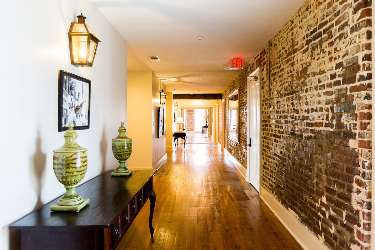A long hallway showcases a combination of modern and historic elements, with exposed brick walls and warm wooden flooring. Two decorative green jars sit on a dark wooden console table. Natural light is seen flowing through large windows at the far end of the corridor.