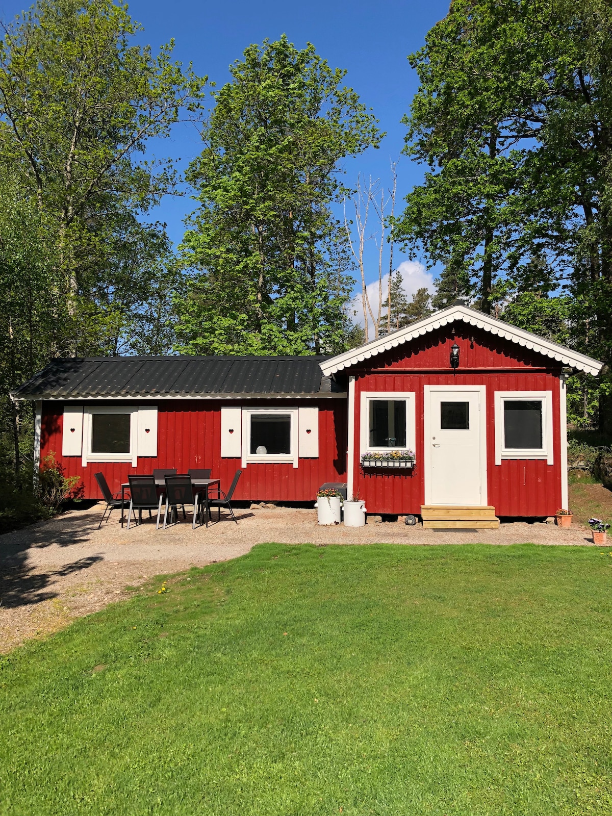 A charming red cabin is surrounded by lush greenery and features a well-maintained lawn. The exterior showcases a combination of a gabled roof and a small porch with steps leading up. A set of black outdoor furniture is placed on the gravel path in front.