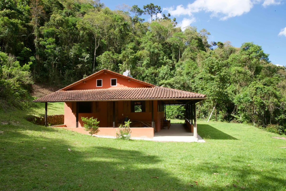 A spacious mountain house is set within lush greenery, featuring a traditional tiled roof and wide porch. The exterior is painted in warm tones, complementing the natural surroundings. The well-kept lawn provides a tranquil space for relaxation, framed by dense trees in the background.