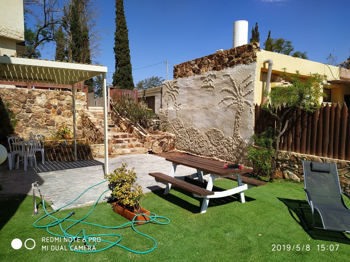 A spacious yard area is displayed, featuring a picnic table and chairs, surrounded by meticulously maintained grass. A stone wall with a palm tree design adds texture to the background. Lush greenery enhances the natural setting, with a few garden elements visible.