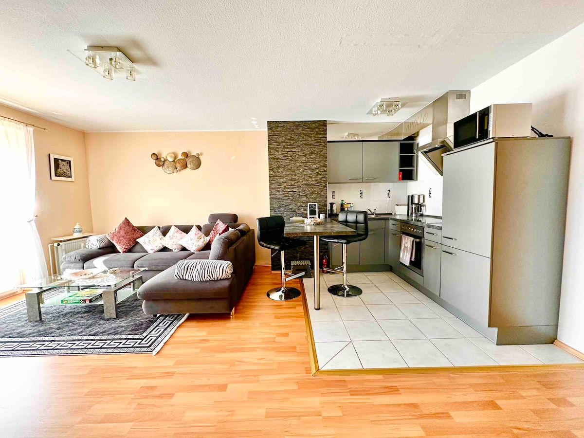 An open-plan living area features a gray sectional sofa adorned with decorative cushions. A dining table and chairs are positioned adjacent to a modern kitchen equipped with stainless steel appliances. Natural light filters through large windows, enhancing the bright and welcoming ambiance.