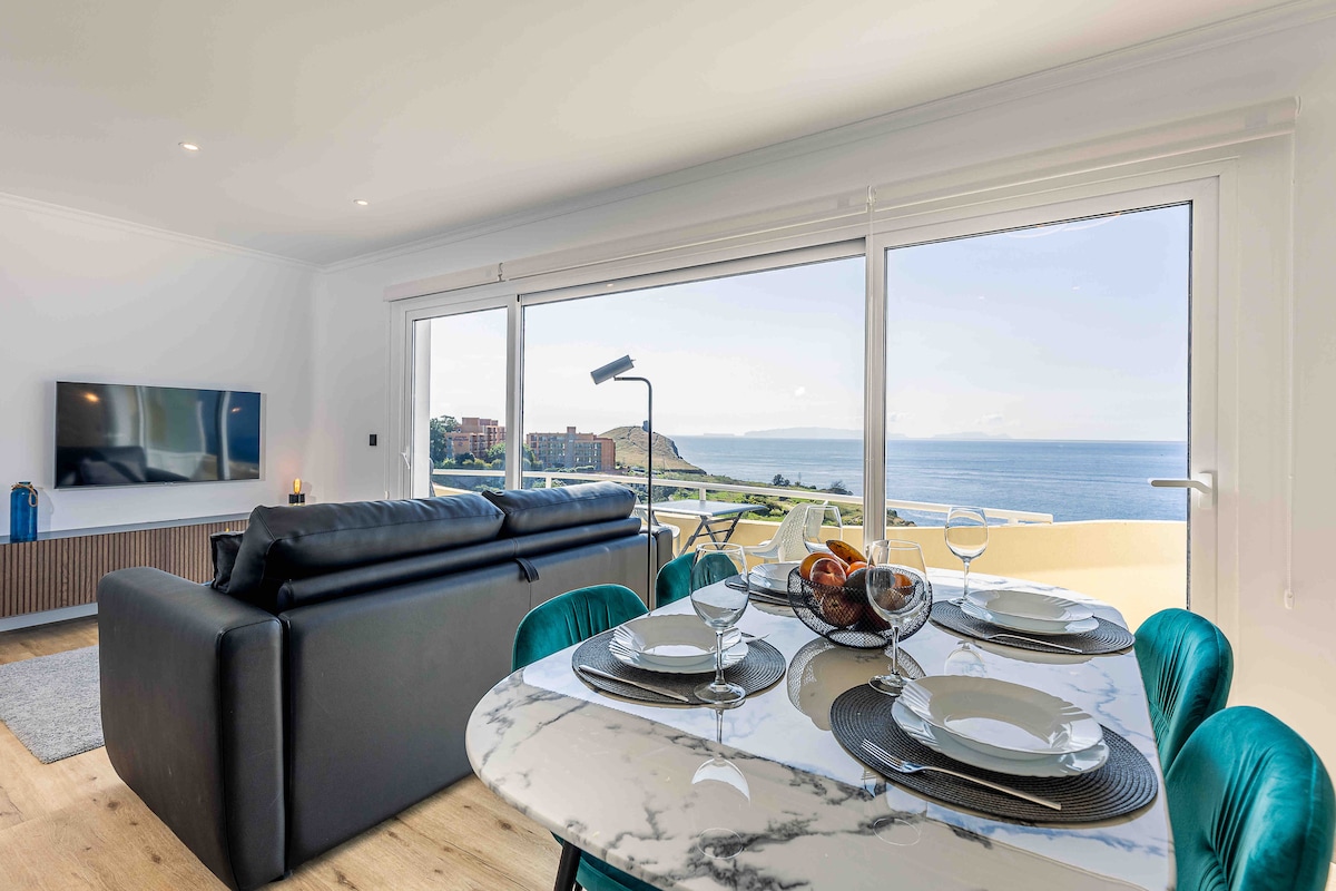 A modern living area is featured, showcasing a sleek black sofa positioned opposite a flat-screen TV. A marble dining table set for four is visible, with beautiful ocean views seen through expansive glass doors that open to a terrace.