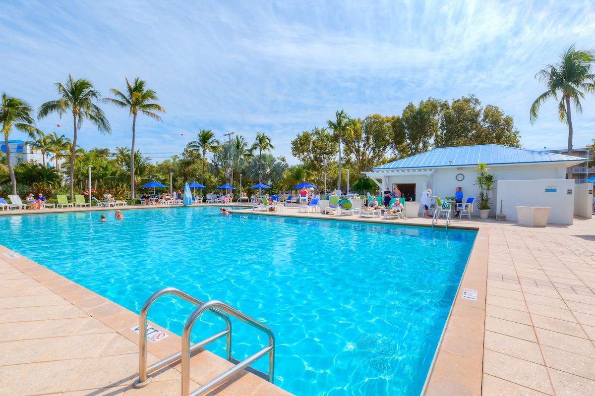A large, inviting swimming pool is surrounded by palm trees, lounge chairs, and umbrellas. The crystal-clear water reflects the blue sky, enhancing the tropical ambiance. A adjacent poolside cabana bar is visible in the background, providing a space for relaxation and refreshments.