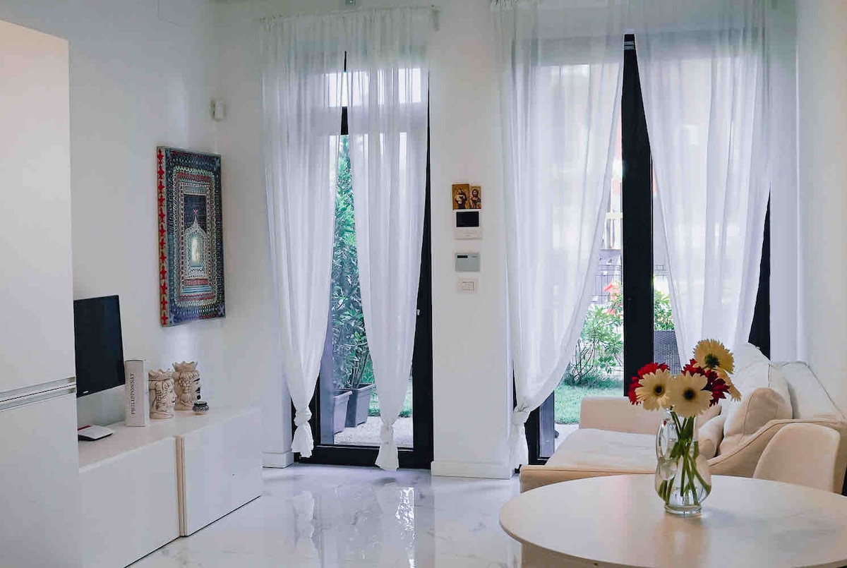 The living area features a light, neutral colour palette with large windows adorned with sheer white curtains. A cozy sofa is positioned beside a round dining table, complementing the bright and airy atmosphere. A vase filled with flowers adds a touch of colour to the space.