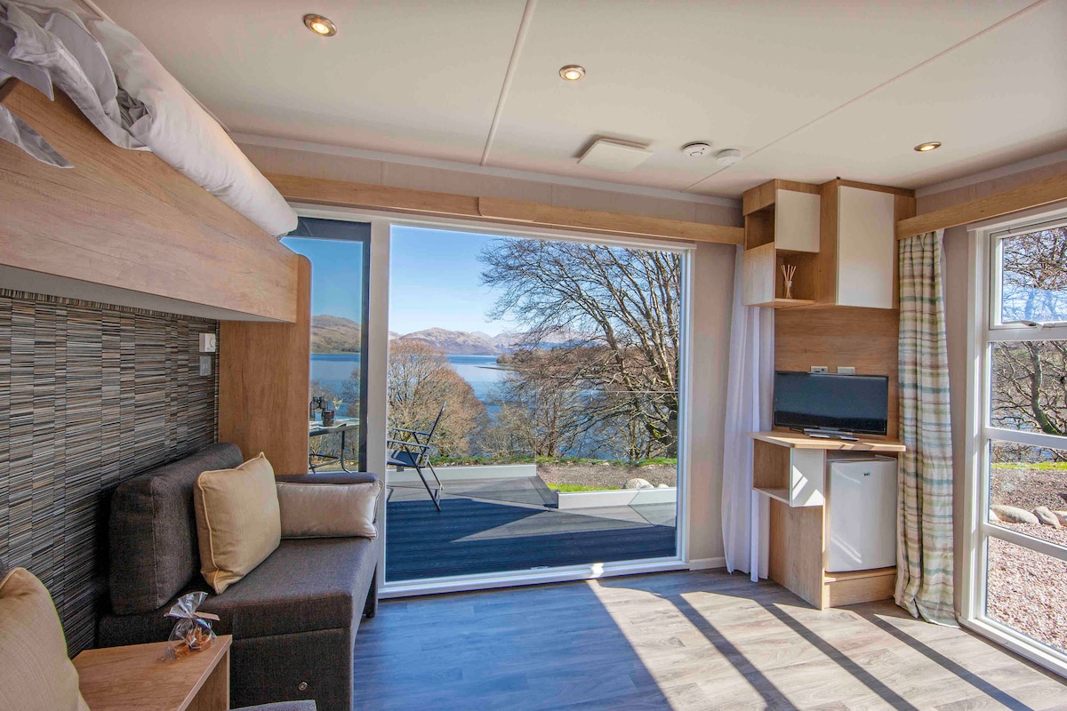 A modern interior space features a cozy seating area with a dark sofa and light-colored cushions. Large glass doors provide a view of Loch Etive, with serene waters and distant hills visible outside. Natural light enhances the warm wooden accents throughout the room.