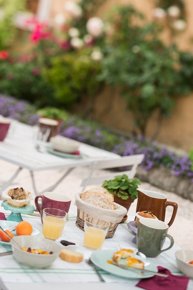 A charming outdoor breakfast setting features a wooden table adorned with plates of food, glasses of juice, and cups of coffee. Freshly baked bread is displayed in a basket, surrounded by colorful flowers and greenery that enhance the tranquil atmosphere.
