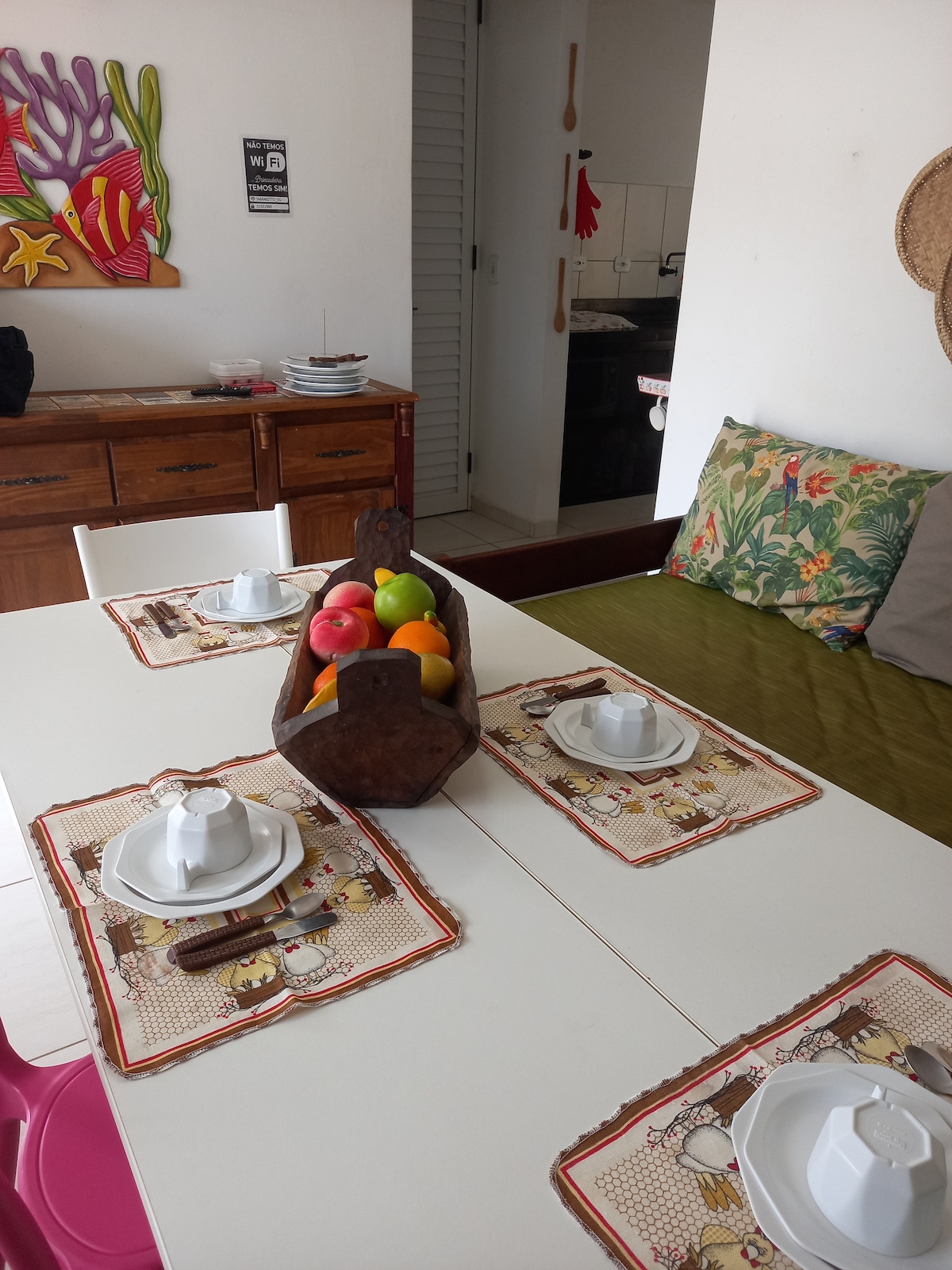 A dining area is presented with a white table set for four, featuring plates and cutlery. A wooden fruit bowl filled with colorful apples and oranges serves as a centerpiece. Vibrant artwork adorns the wall, while a cozy green seating area is visible nearby.