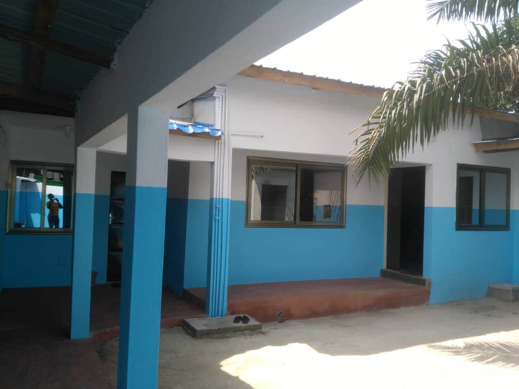 The exterior view showcases a single-story building featuring a distinct blue and white color scheme. Windows with dark frames provide natural light to the interior spaces. A paved pathway leads towards the entrance, bordered by sandy areas and greenery from nearby palm trees.