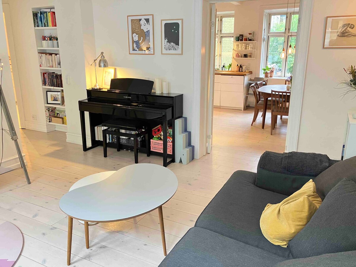 An airy living area is highlighted by a modern gray sofa and a low coffee table with wooden legs. A black piano sits against the wall, accompanied by framed art. The space flows into a bright kitchen and dining area, visible through an open doorway.