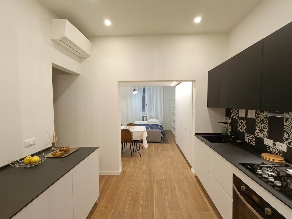 The modern kitchen displays sleek black cabinets and a dark countertop. A small dining area is visible through an open doorway, featuring a table and chairs. Light-colored walls enhance the spacious feel, while subtle decorative tiles add character.