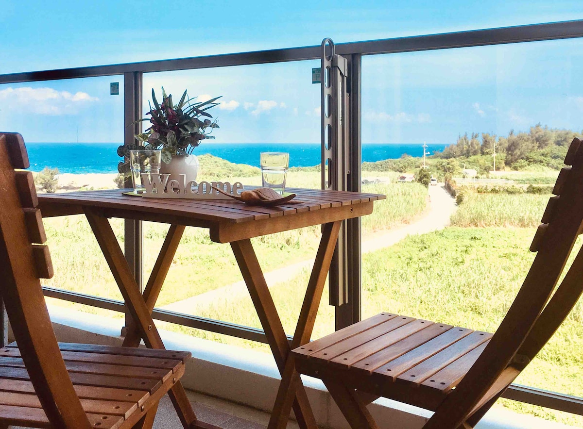 A wooden table set for two is positioned on a private balcony, offering an ocean view. The horizon displays a vibrant blue sky meeting the sea, while greenery of beach vegetation can be seen below. A small potted plant decorates the table, creating a welcoming ambiance.