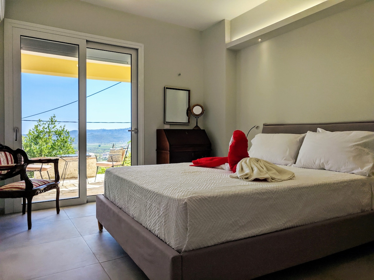 A spacious bedroom features a large bed with soft linens and a striking red pillow. Natural light fills the room from glass doors that open to a balcony, revealing panoramic views of green plains and the Ionian Sea. A chair and a wooden dresser complement the decor.