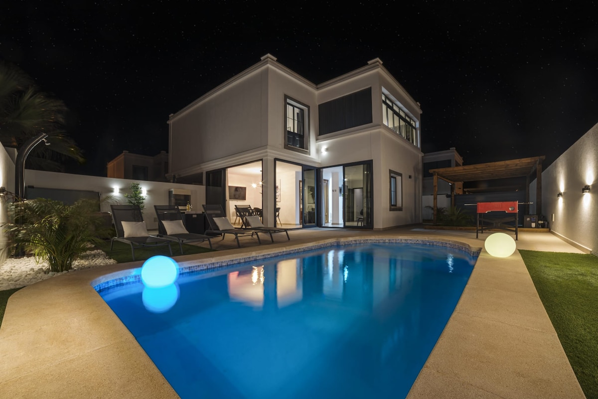 A private outdoor area features a heated swimming pool with illuminated accents, reflecting the night sky. The two-story villa, with large glass doors, offers a view into the well-lit living space. Comfortable lounge chairs are arranged poolside, surrounded by lush greenery.