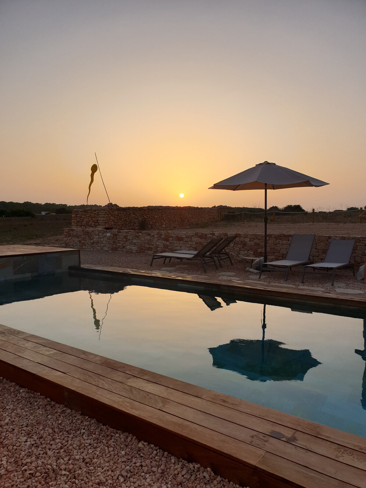 The tranquil pool reflects the soft colors of a sunset, with an umbrella providing shade over the lounge chairs. Surrounding stone walls add rustic charm, while the open landscape in the background enhances the serene atmosphere.