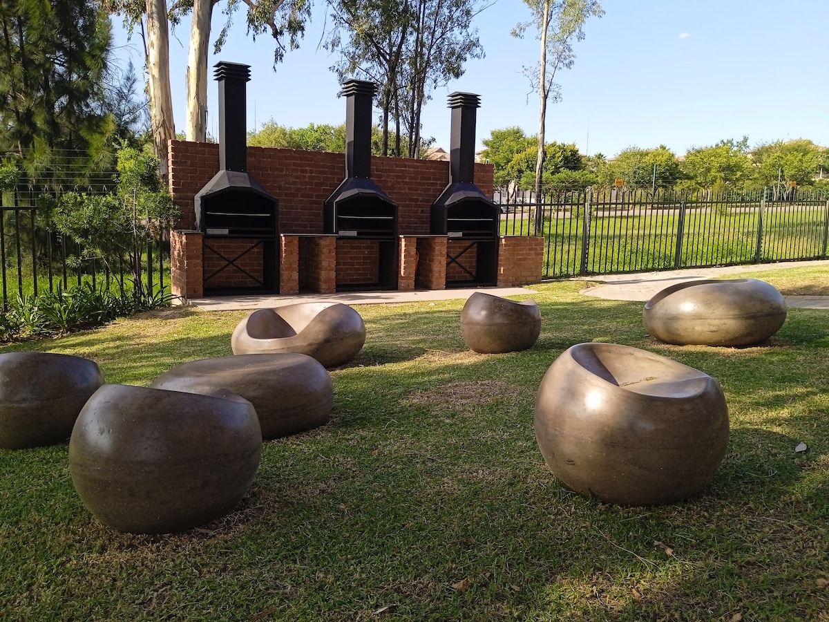A landscaped area features sculptural stones arranged on the grass, providing unique seating. In the background, three charcoal grills are placed within a brick structure, surrounded by a fenced garden, under the shade of trees.