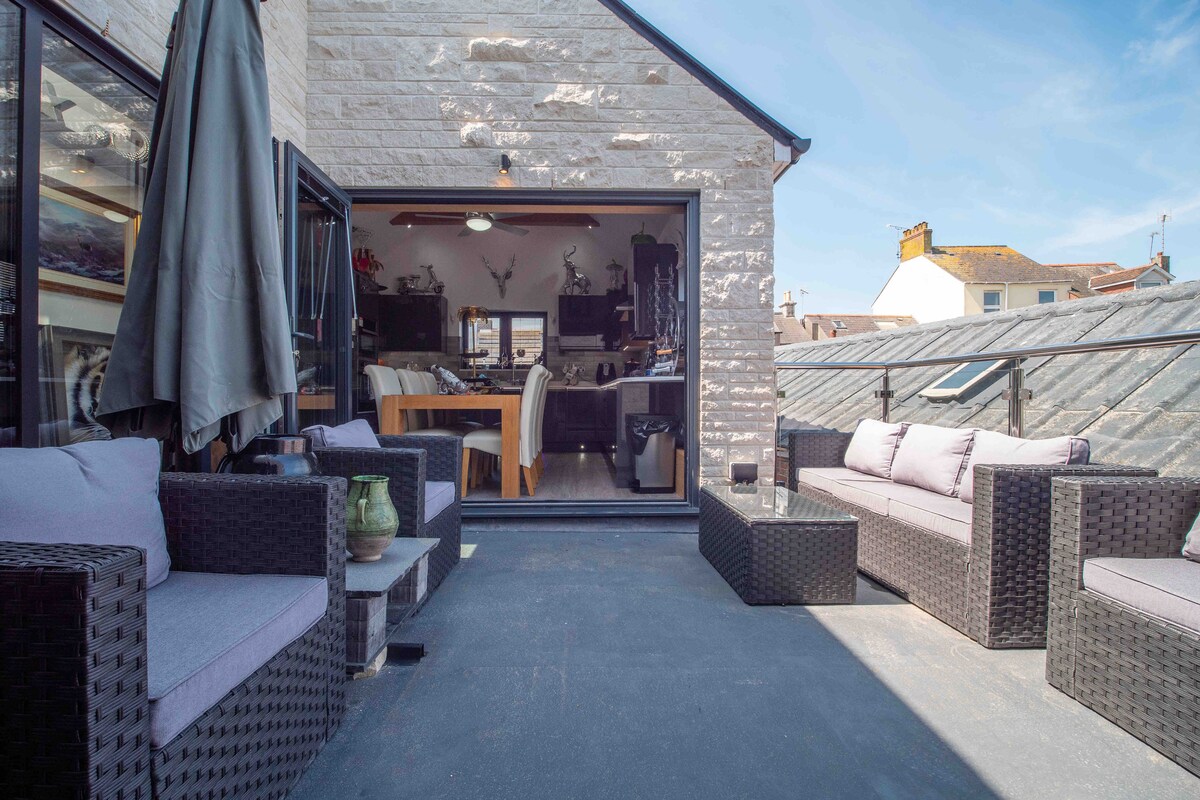 An outdoor mezzanine area is presented, featuring comfortable seating arranged around a central coffee table. Bi-folding doors provide a seamless connection to the interior space, where a dining area can be seen. Natural light fills the area, accentuating the stone exterior.