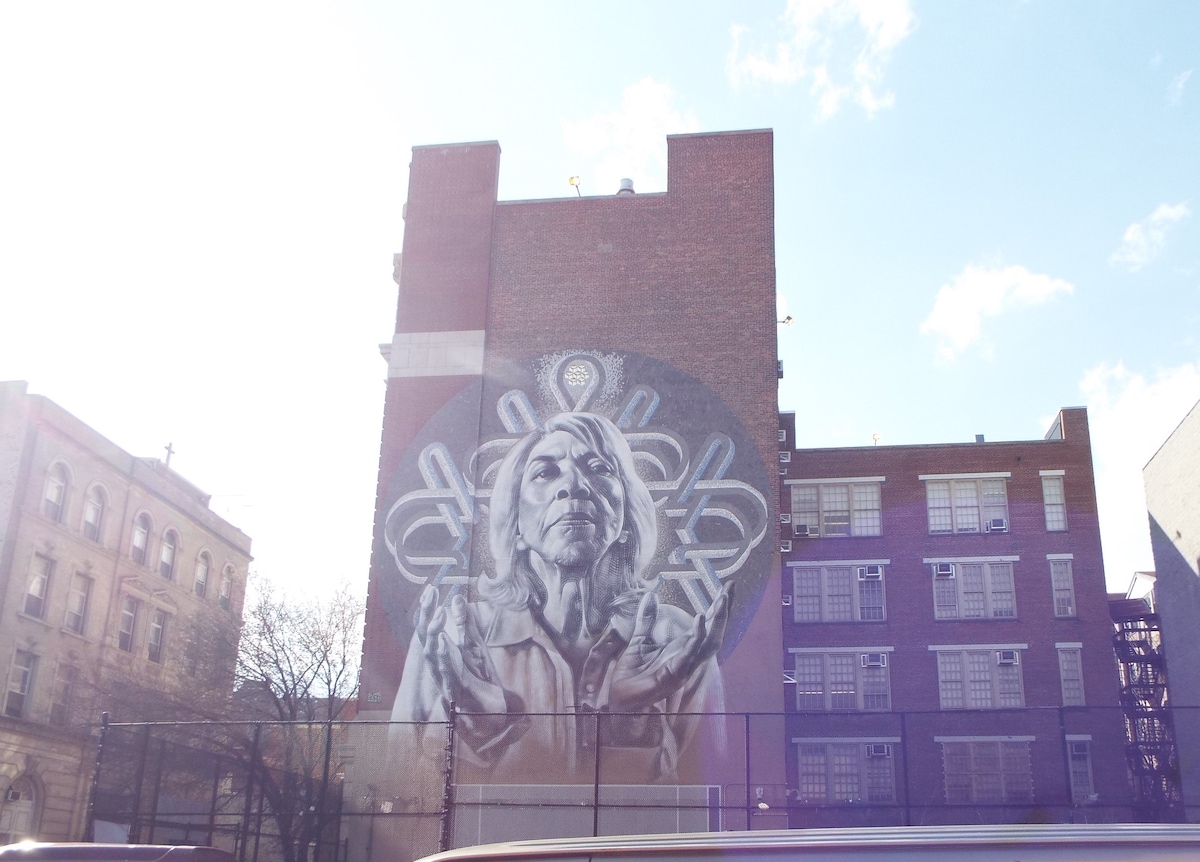 A vibrant mural featuring intricate hand designs is displayed on a brick building. Sunlight illuminates the artwork, creating a striking contrast against the surrounding urban architecture. Nearby structures include residential buildings with multiple windows, emphasizing the community's artistic character.