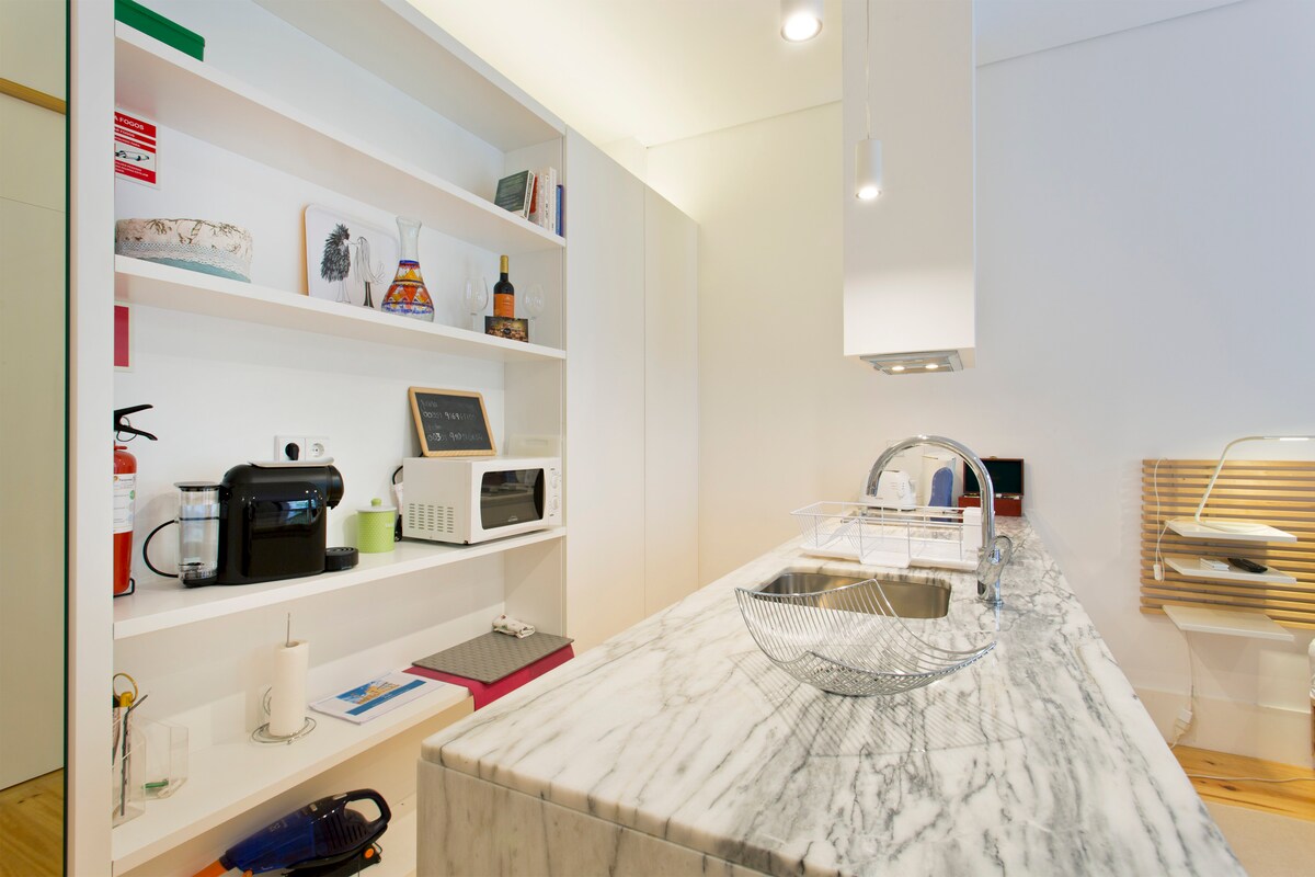 The kitchen area features a sleek marble countertop with a stainless-steel sink. Above, open shelving displays various kitchen items, including a coffee maker, microwave, and decorative bottles. A modern exhaust hood hangs overhead, illuminating the workspace with natural light.