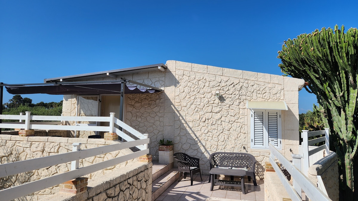 The exterior of the house showcases a light stone facade, featuring outdoor seating arranged under a shaded area. A cactus plant provides green accents, while a clear blue sky serves as a backdrop. The entrance is accessible via a few steps leading to a raised terrace area.