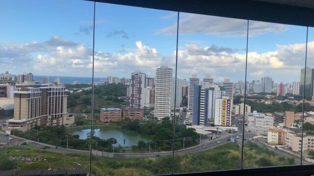 A panoramic view of the cityscape showcases high-rise buildings and greenery, with a scenic lake visible in the foreground. The ocean is seen on the horizon under a partly cloudy sky, which enhances the sense of openness in the landscape.