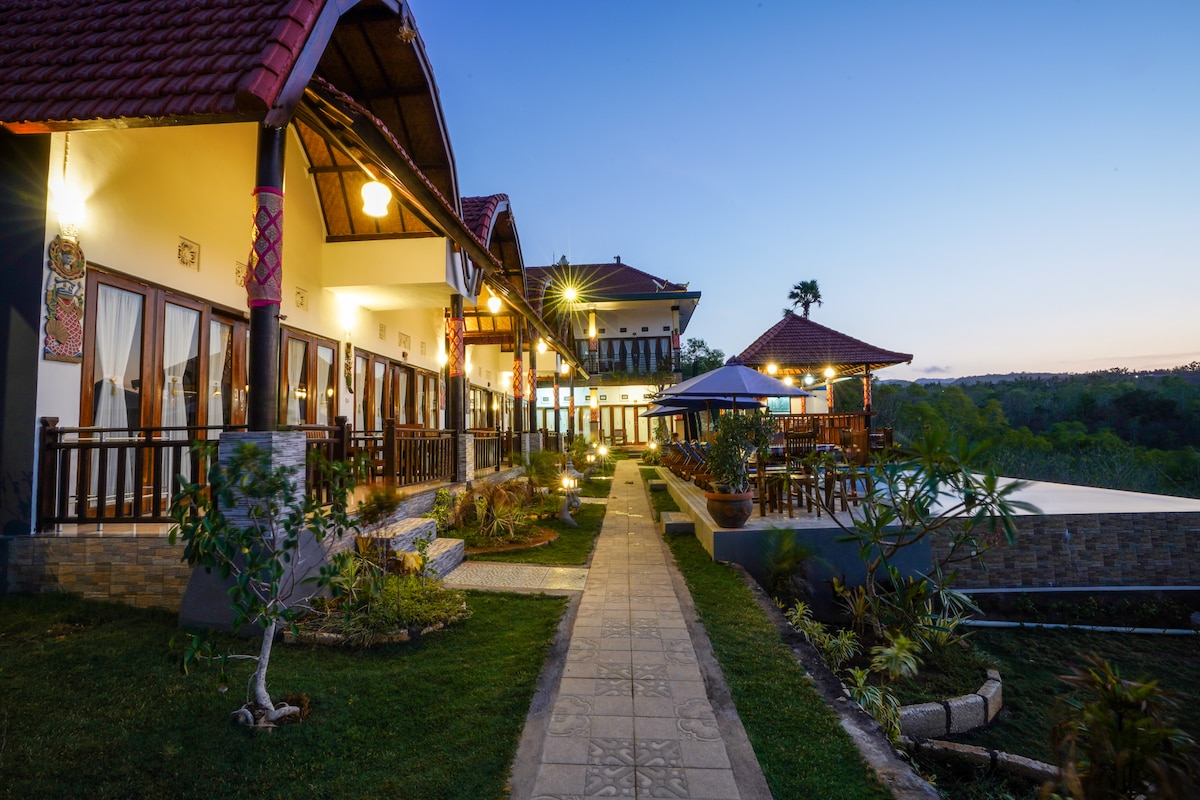 A pathway leads through a well-maintained garden, bordered by villas displaying traditional architecture. Illuminated in soft lighting, the structures feature multiple balconies with glass doors. Lush greenery complements the serene ambiance, and the sunlight begins to fade, creating a peaceful evening atmosphere.