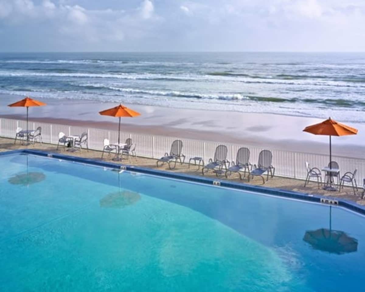 An outdoor pool area offers a spacious swimming area, flanked by lounge chairs under orange umbrellas. The ocean waves gently lap against the shore beyond, providing a serene backdrop to this relaxing space.