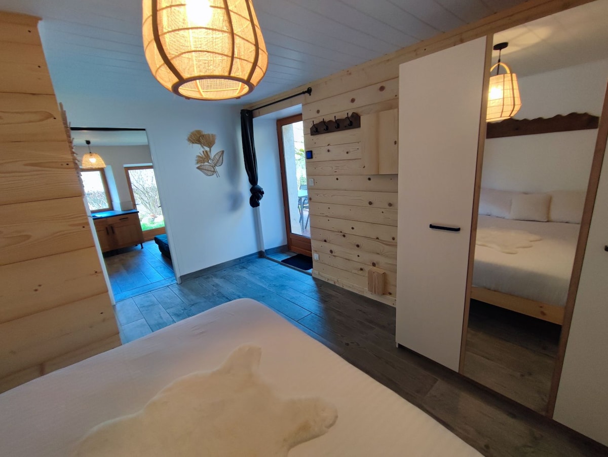 A serene bedroom space features a spacious bed with a soft, light-coloured accent rug. Natural wood paneling enhances the warmth of the room. A doorway leads to an outdoor area, complemented by soft lighting from pendant fixtures.