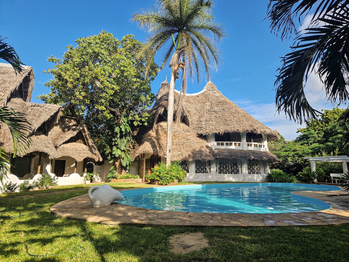 The villa features a charming thatched roof, complemented by lush greenery and palm trees. A serene swimming pool is positioned in the foreground, surrounded by a spacious garden area, creating a relaxing outdoor environment for leisure and enjoyment.