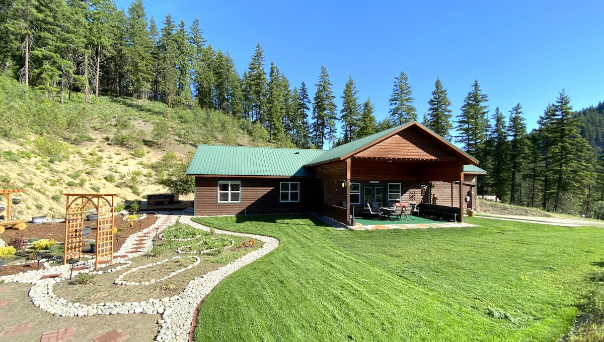 A spacious lodge is set amidst tall trees, featuring a green metal roof and a large covered porch. The manicured lawn is accented by a stone pathway and flower beds, inviting guests to enjoy the serene outdoor space.