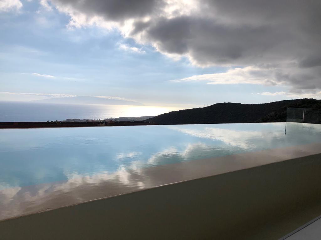 An infinity pool extends towards the horizon, reflecting the sky and surrounding landscape. Gentle waves of water blend with the serene ocean, while clouds drift overhead. The view includes hills in the background, creating a seamless connection between land and sea.