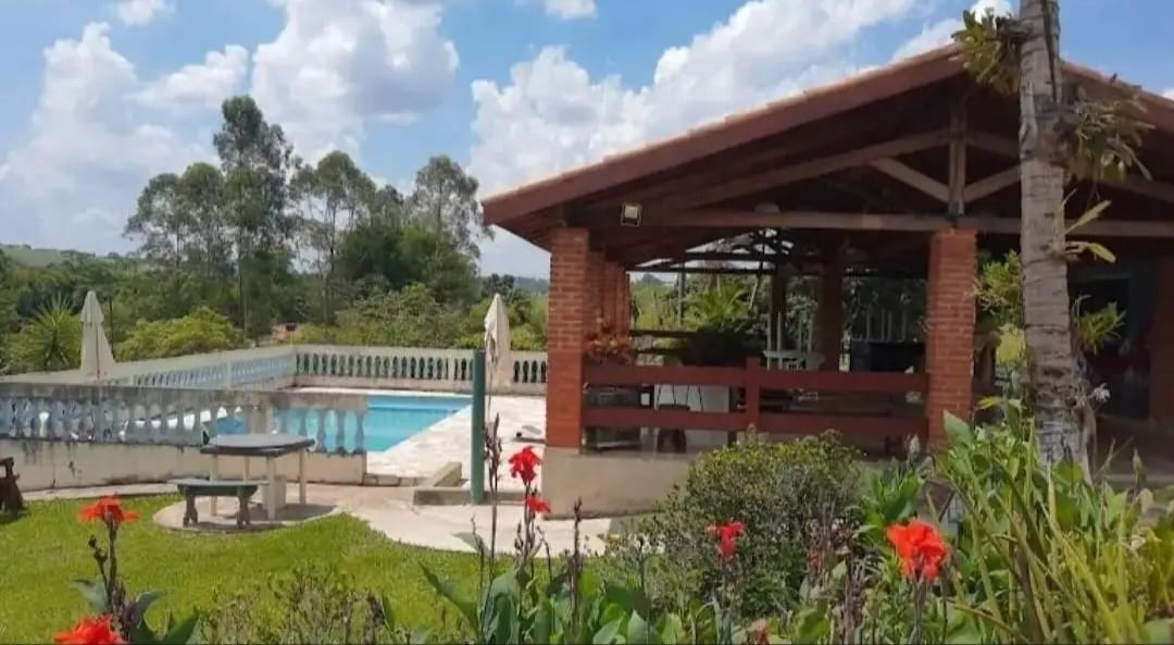 An outdoor area features a brick pavilion with a shaded terrace overlooking a large swimming pool. Lush greenery surrounds the space, with vibrant flowers adding color. The serene landscape includes trees in the background, enhancing the overall natural setting.