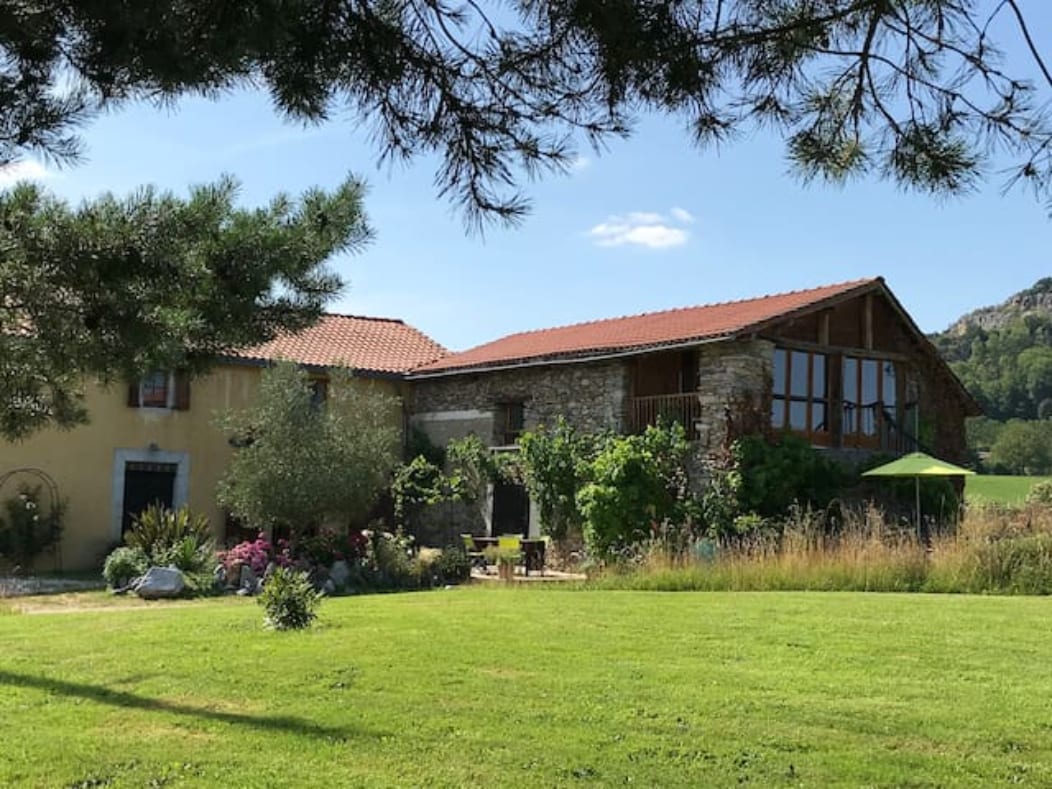 A charming gîte is nestled in a lush green field, surrounded by trees and colorful flowers. The traditional stone structure features a large, open terrace with wooden beams and a sloped roof, inviting guests to enjoy the serene countryside setting.