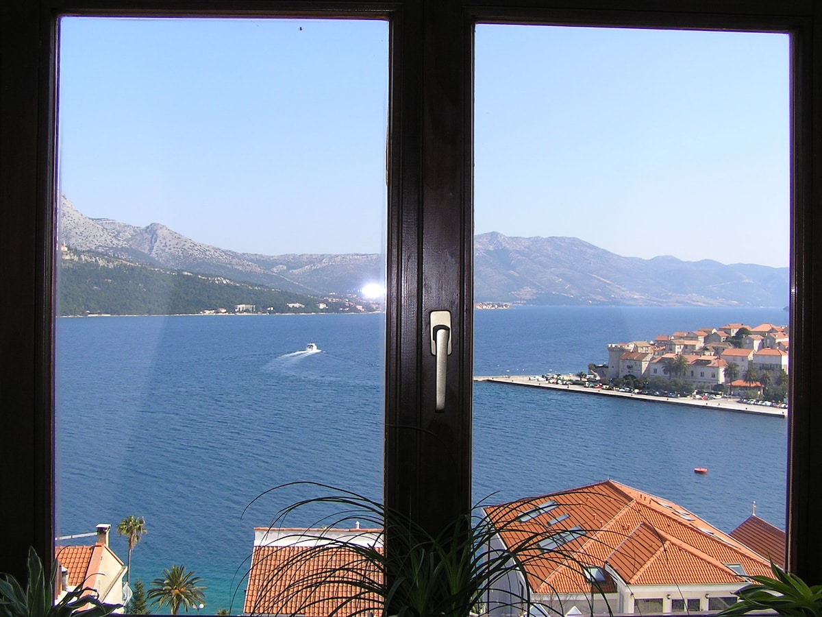 A panoramic view of the Pelješac channel showcases the serene blue waters and mountains in the background. The window frame highlights the coastal town with terracotta rooftops and a distant boat on the water, creating a peaceful seaside atmosphere.