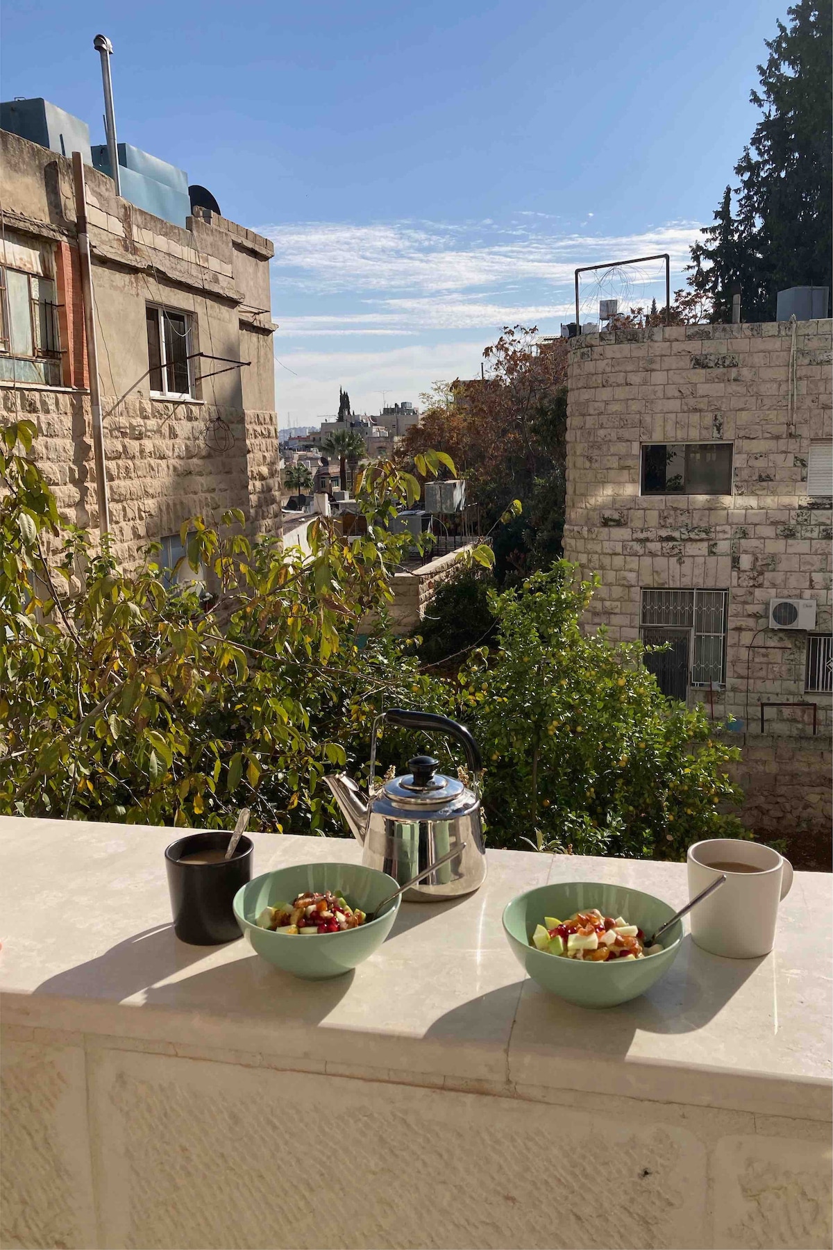 A private balcony view reveals buildings with stone facades and greenery. A kettle and two bowls of salad are placed on the ledge, with a cup beside them. The morning sunlight illuminates the scene, highlighting the textures of the surrounding architecture.