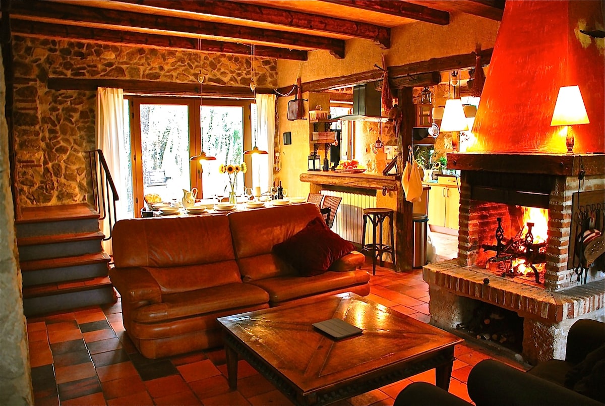 A spacious living area features a large leather sofa facing a warm fireplace. The room is accented with exposed wooden beams and stone walls. A dining table can be seen in the background, illuminated by soft lighting, with a view of the outdoors through large glass doors.