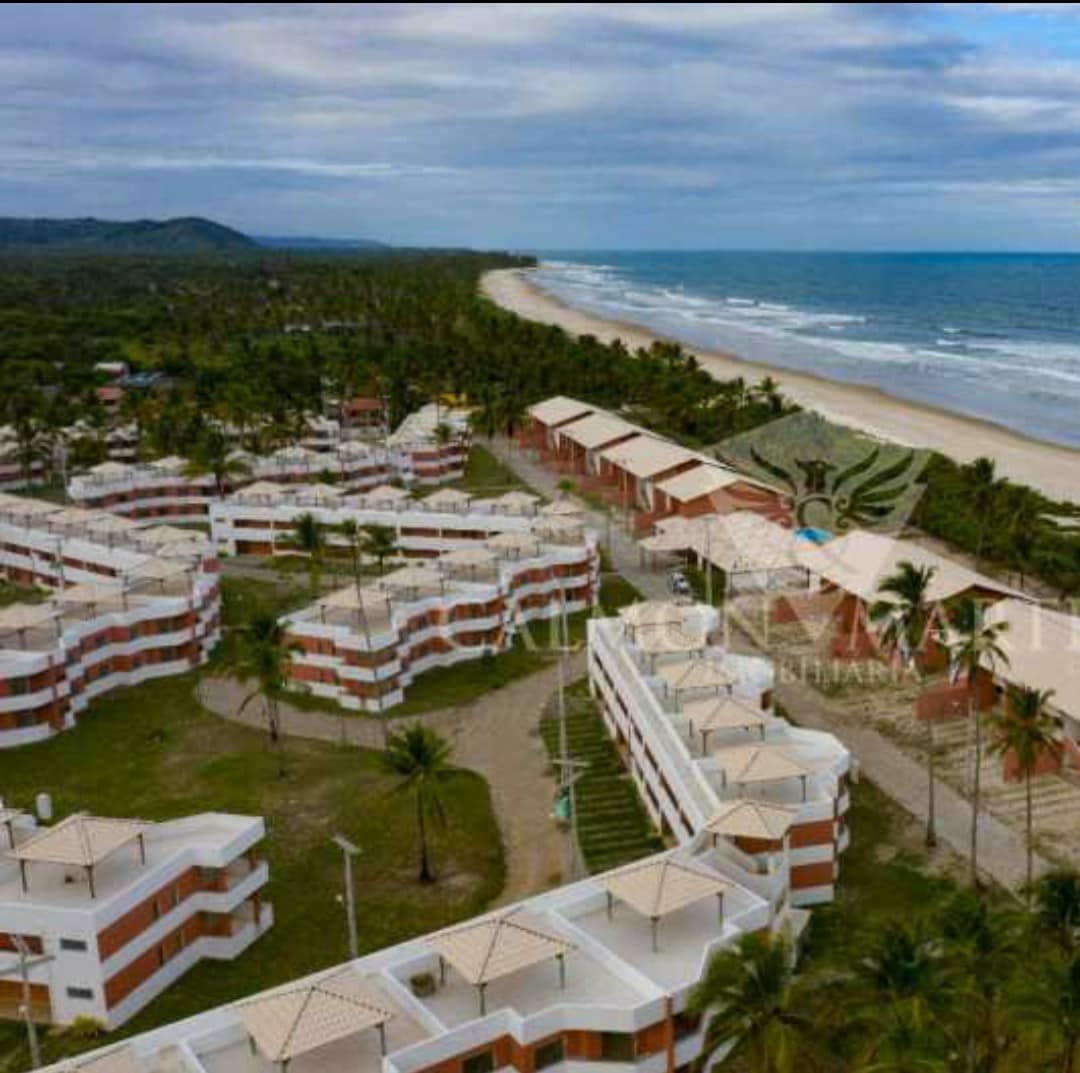 An aerial view captures a resort layout surrounded by palm trees, with multiple white and red building clusters. A serene beach is visible in the background, featuring gentle waves meeting the shoreline, and the landscape is complemented by lush greenery.