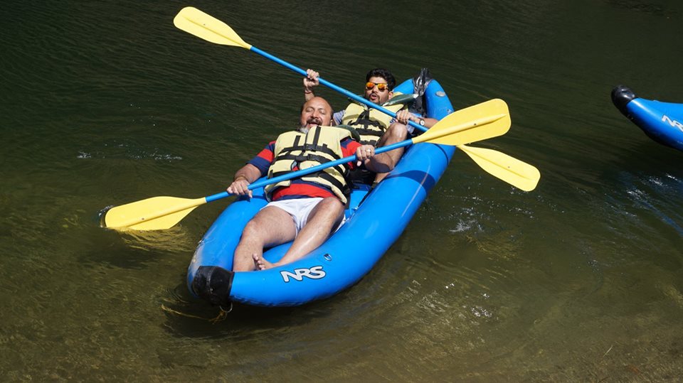 A bright blue kayak is visible on calm waters, occupied by two individuals wearing yellow life vests. Each person holds a yellow paddle, creating a contrast against the surrounding natural scenery. The clear water reflects the kayak, enhancing the serene environment.