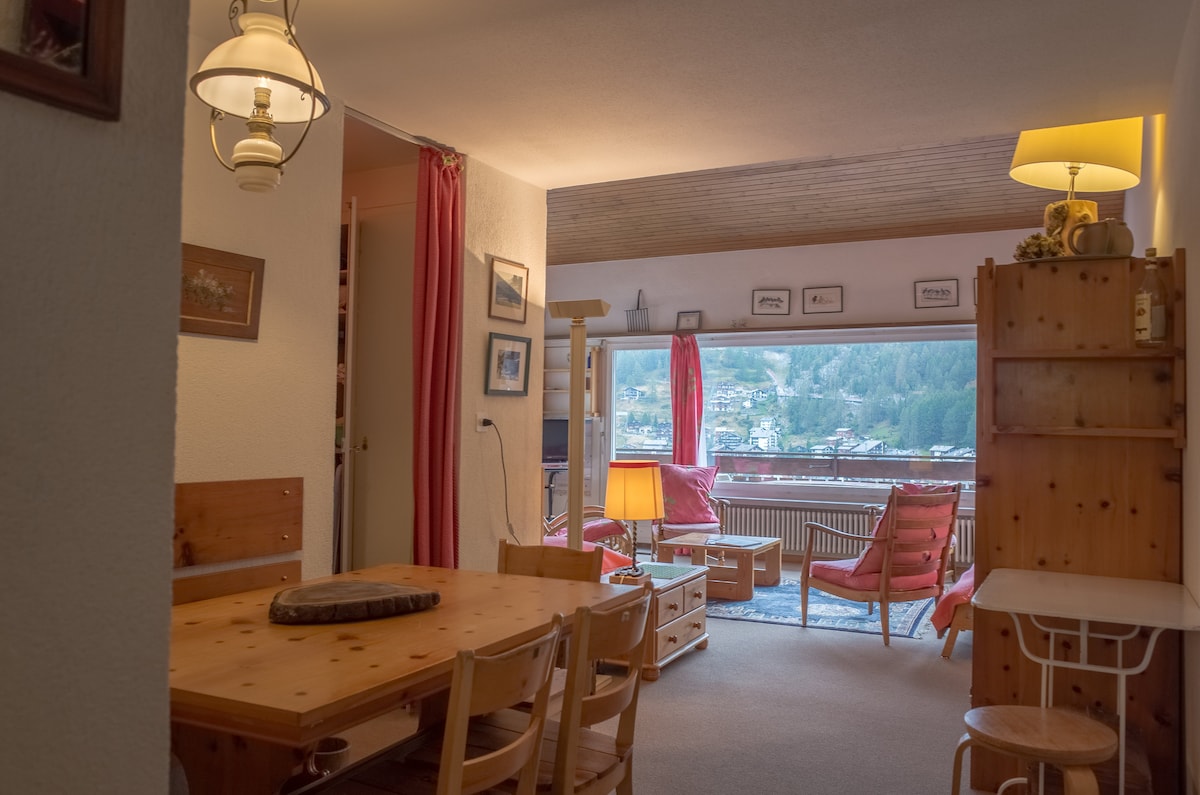 The living area features a wooden dining table surrounded by chairs, with a cozy seating arrangement in front of large windows offering mountain views. Light fixtures provide warm illumination, while decorative elements and wall art enhance the space's character.