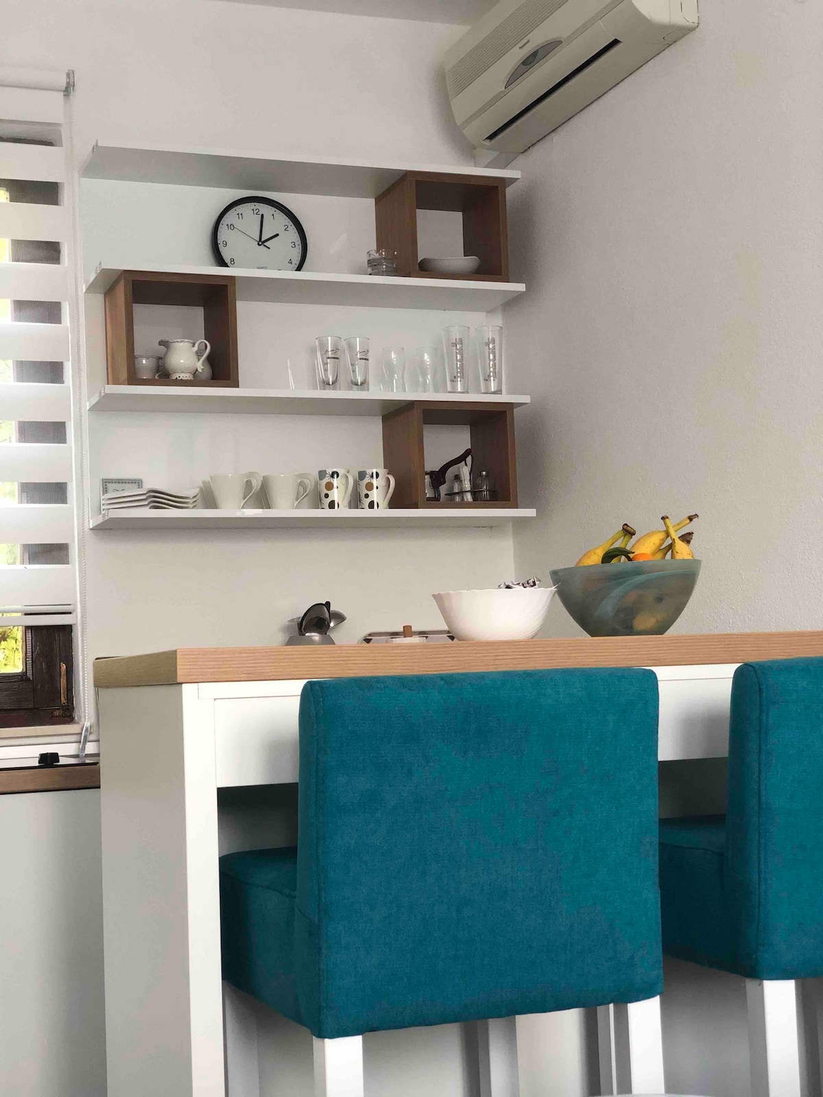 A compact dining area features a white table with two teal stools. A wall-mounted shelf displays glassware and ceramic items. A fruit bowl with bananas sits on the table, while a clock and small decorative pieces adorn the shelf above, providing a functional yet stylish look.